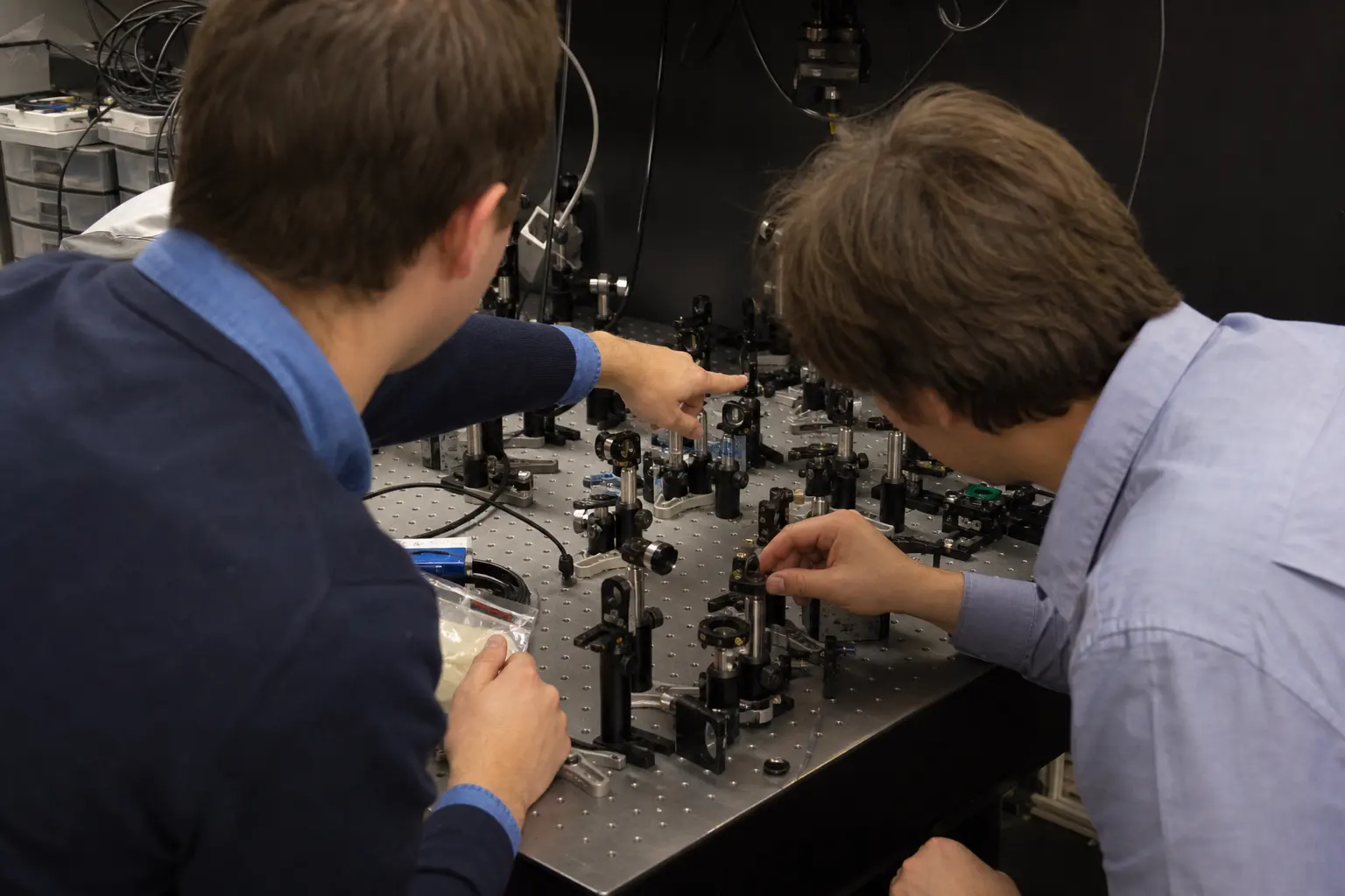 Two engineers working together at an optics bench covered with mounts, mirrors and cabling, seen from behind.